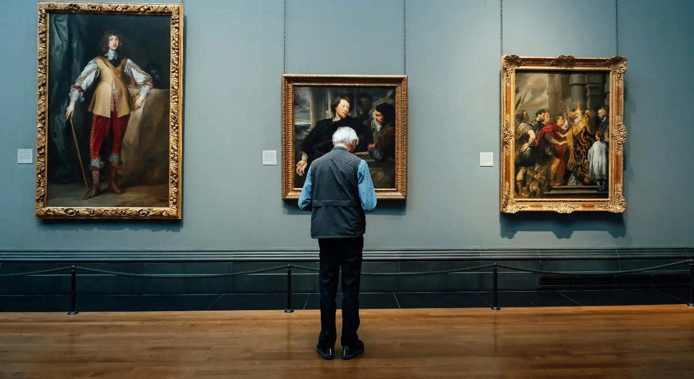 Старац стоји у галерију испред три фотографије окачене на зид.