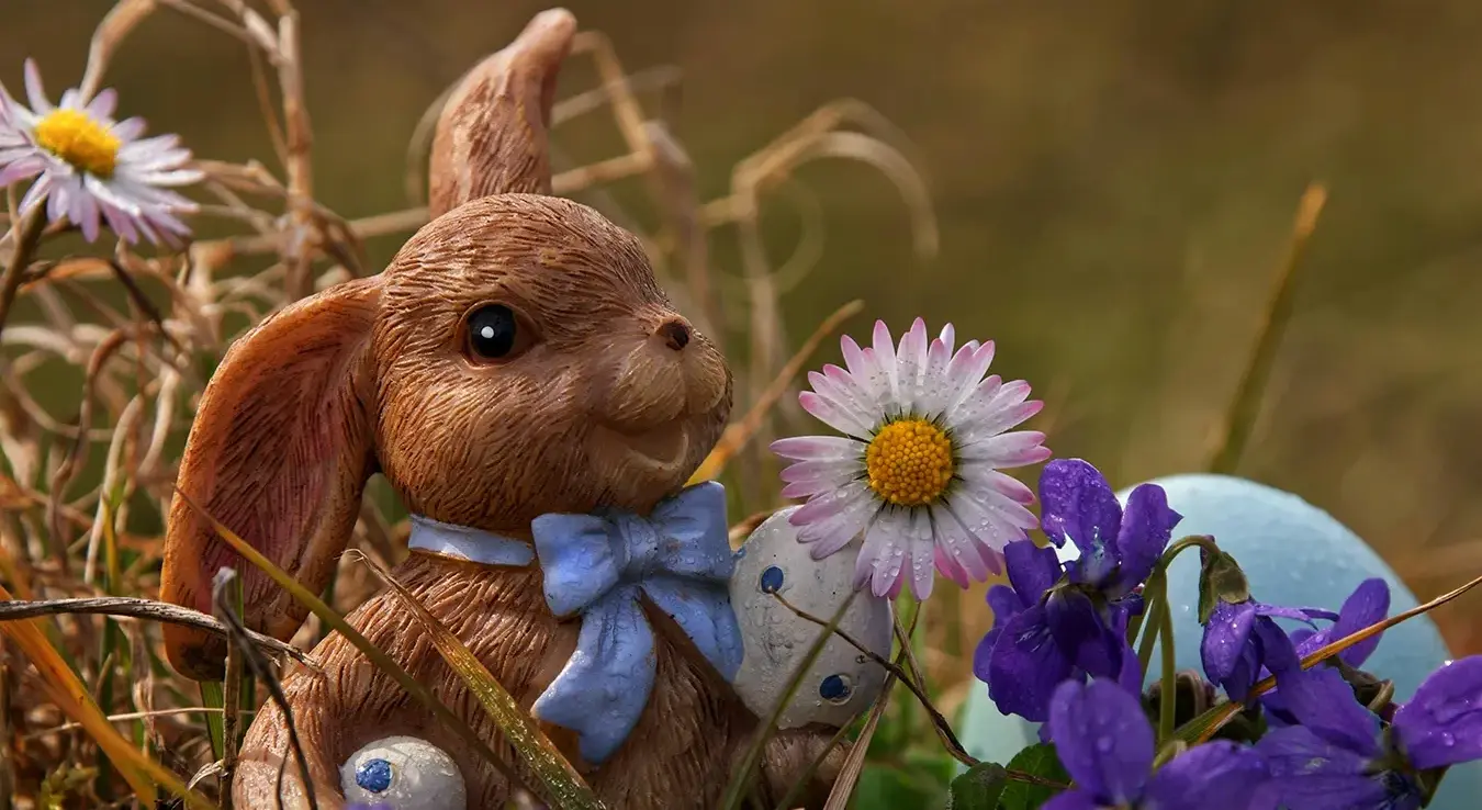 Васкршњи зец са плавом машном и јајима око њега стоји у трави.