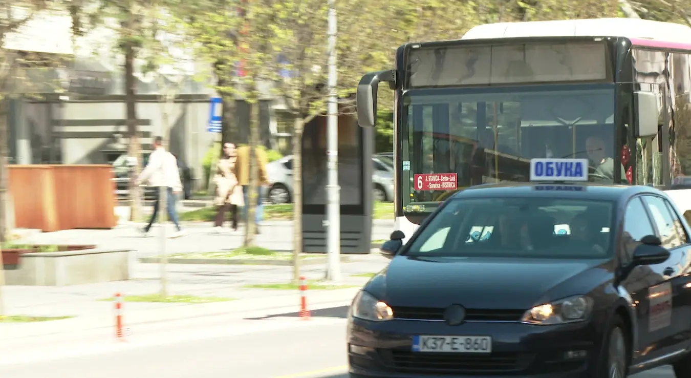 Иза аутомобила налази се бијели градски аутобус. На његовој предњој страни стоји ознака линије број 6, која саобраћа на релацији А.