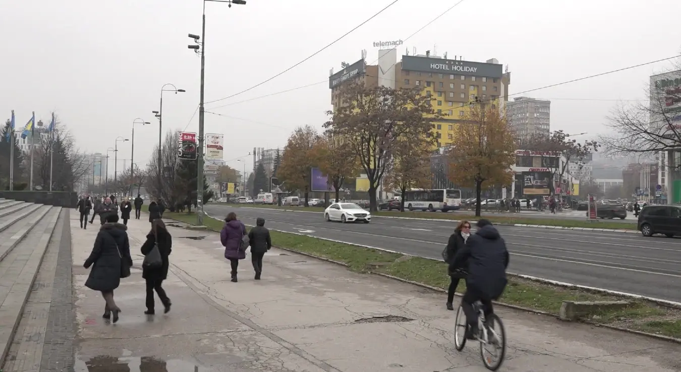 Панорама Сарајева са погледом на хотел Холидеј и главну саобраћајницу. Пролазници на тротоару и бициклиста испред Парламентарне скупштине БиХ током облачног дана.
