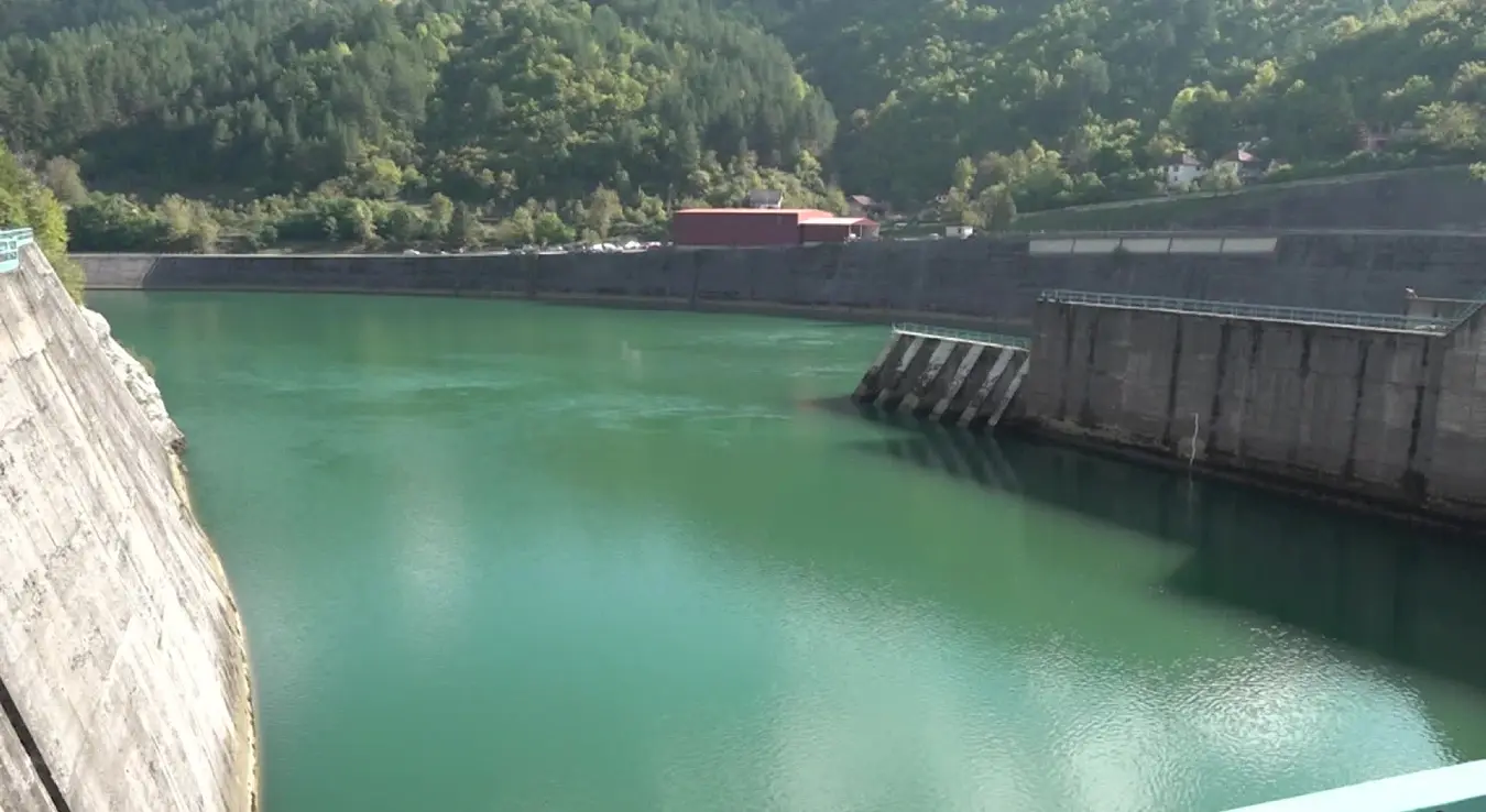 Tок ријеке Дрине испред бетонске бране ХЕ Вишеград. У фокусу су масивни потпорни зидови објекта и дио постројења, док се у позадини виде стрма брда прекривена густом шумом под дневним свјетлом.