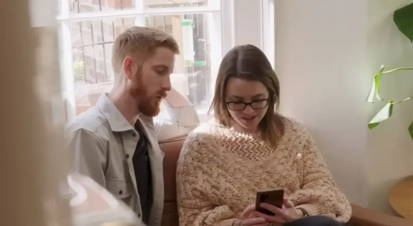 Мушкарац и жена сједе на каучу и гледају у телефон.