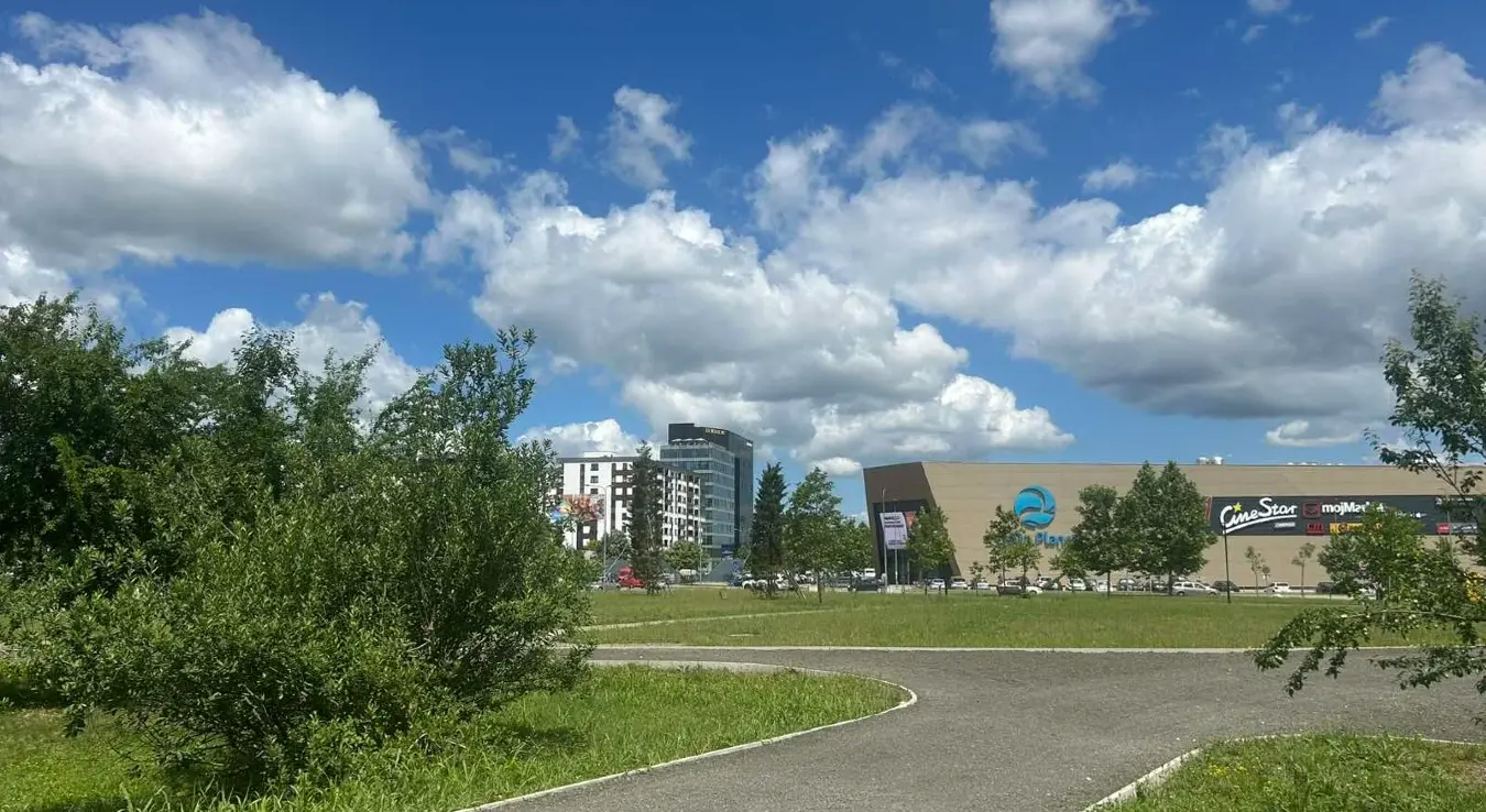 Приказан је сунчан дан у градском парку са асфалтном стазом у предњем плану и тржним центром у позадини.
