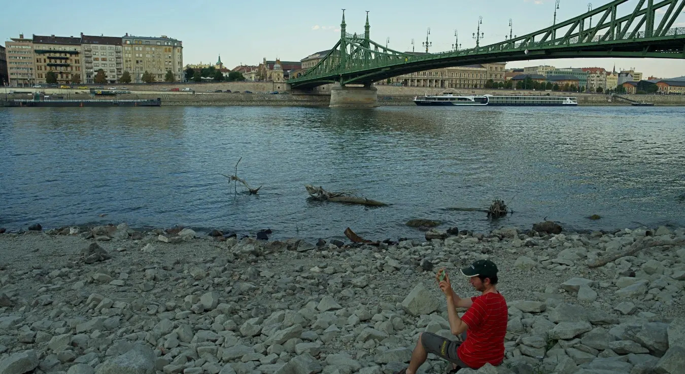 Мушкарац фотографише док сједи на сувој обали Дунава у центру Будимпеште у Мађарској, у сриједу, 5. августа 2026. године, док је ниво ријеке, која протиче кроз 10 европских земаља, опао на до сада незабиљежено низак ниво.