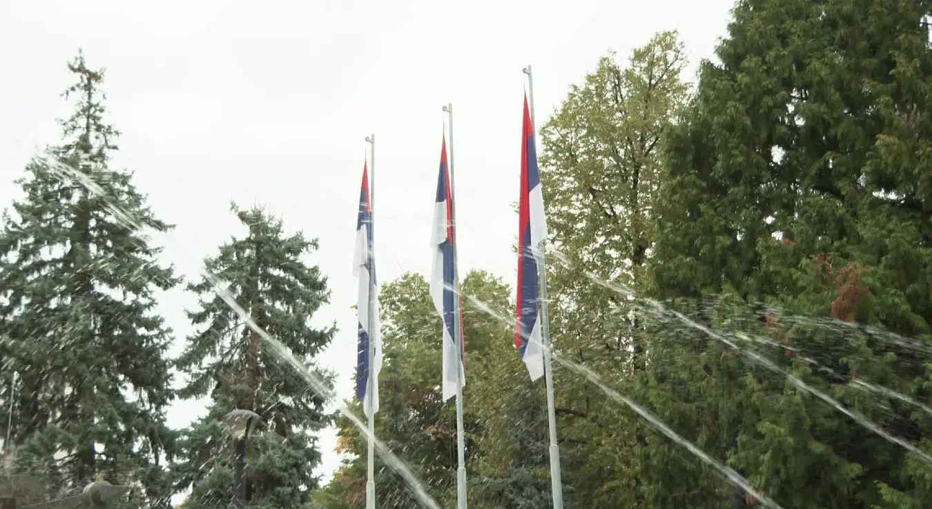 Заставе Републике Српске вијоре се на јарболима испред објекта, док млазеви воде прелазе преко њих и околног дрвећа током заливања зелених површина.