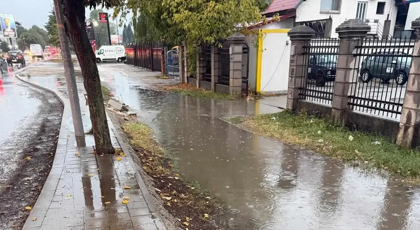Неуређена пјешачка стаза прекривена је водом и блатом, док се на тротоару задржавају велике локве. Овакви услови отежавају кретање пјешака, посебно дјеце, старијих и особа са отежаним кретањем. Фотографија указује на проблеме са проходношћу и одводњом воде након кише.