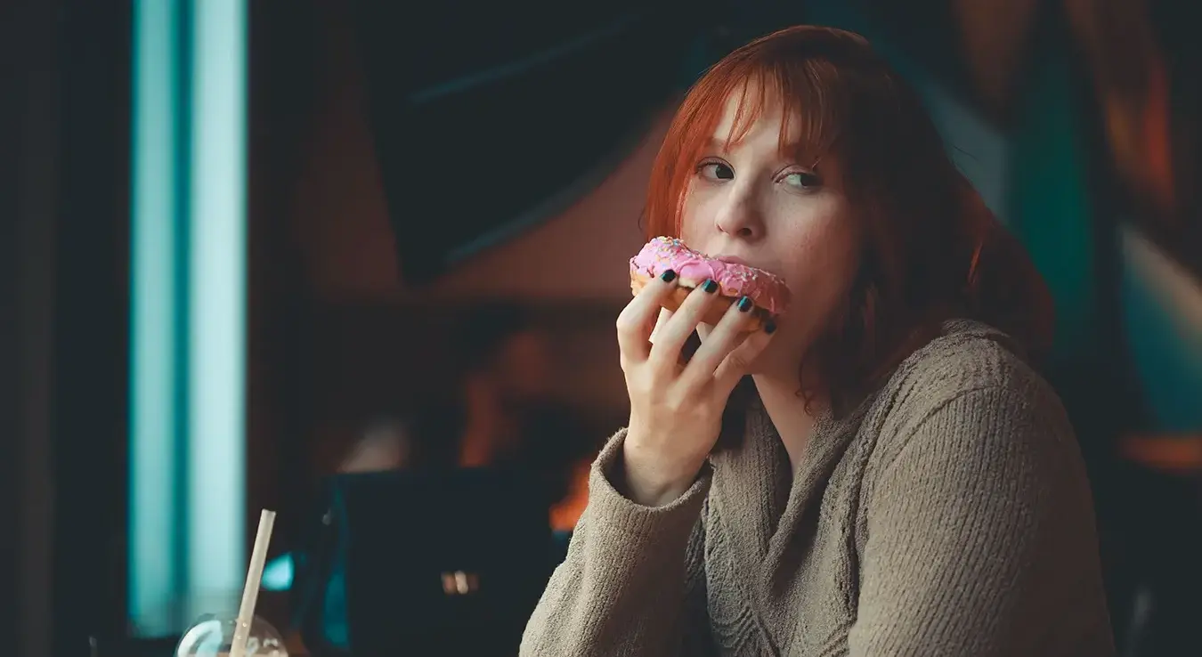 Риђокоса жена ужива у ружичастој крофни у затвореном простору кафића.